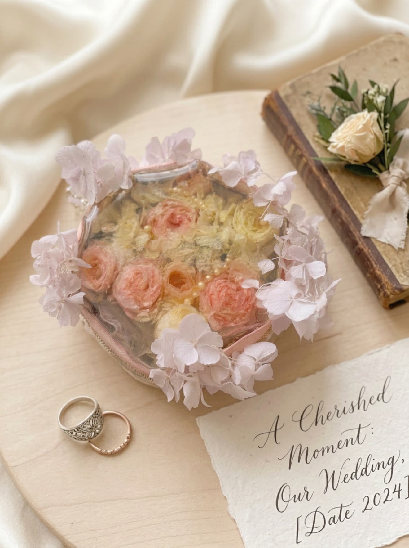 Clear pouch with dried roses, wedding rings, and a handwritten note on a wooden surface.