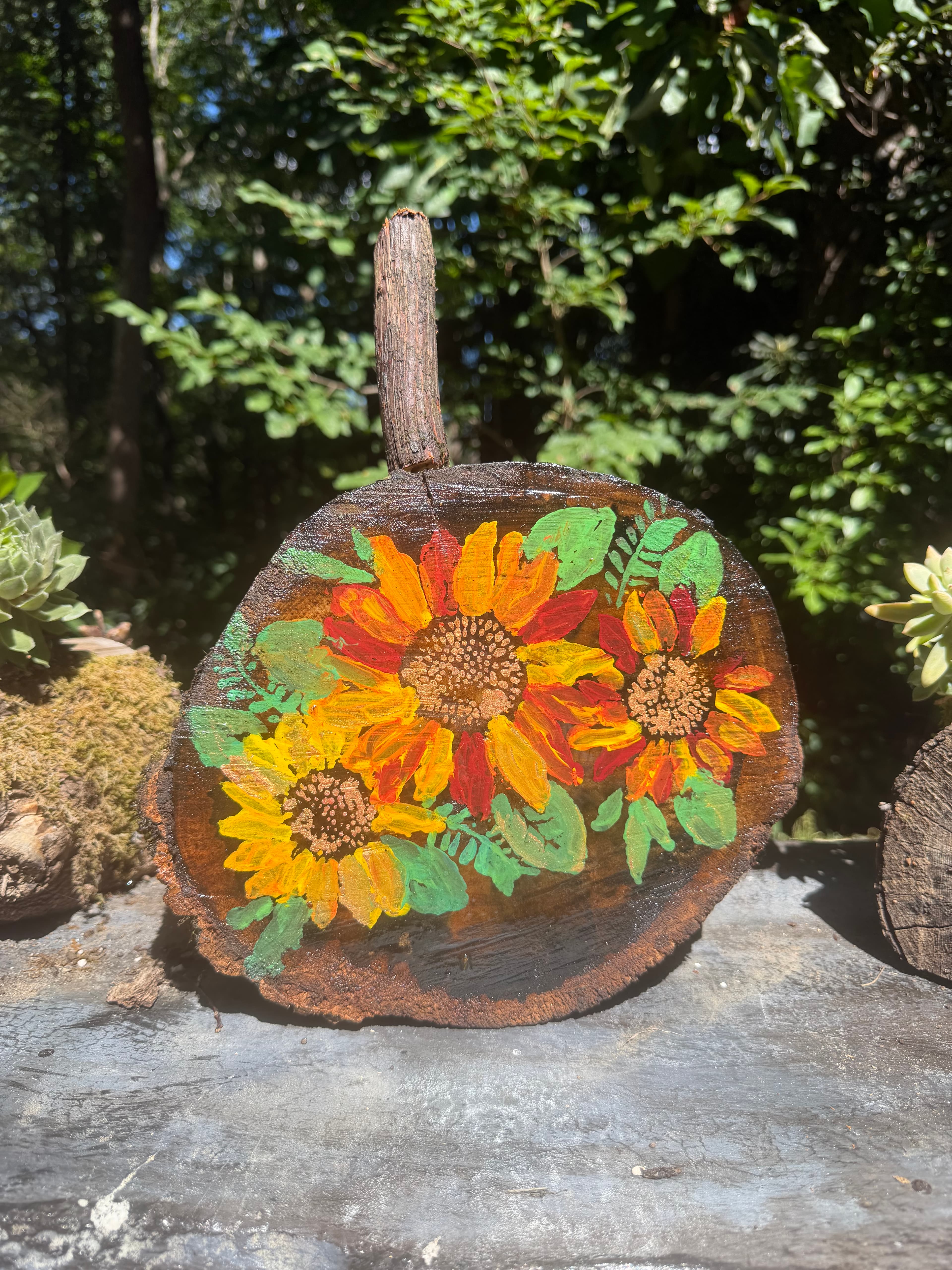Rustic wood slice pumpkin decorated with vibrant hand-painted sunflowers, set against a leafy background.