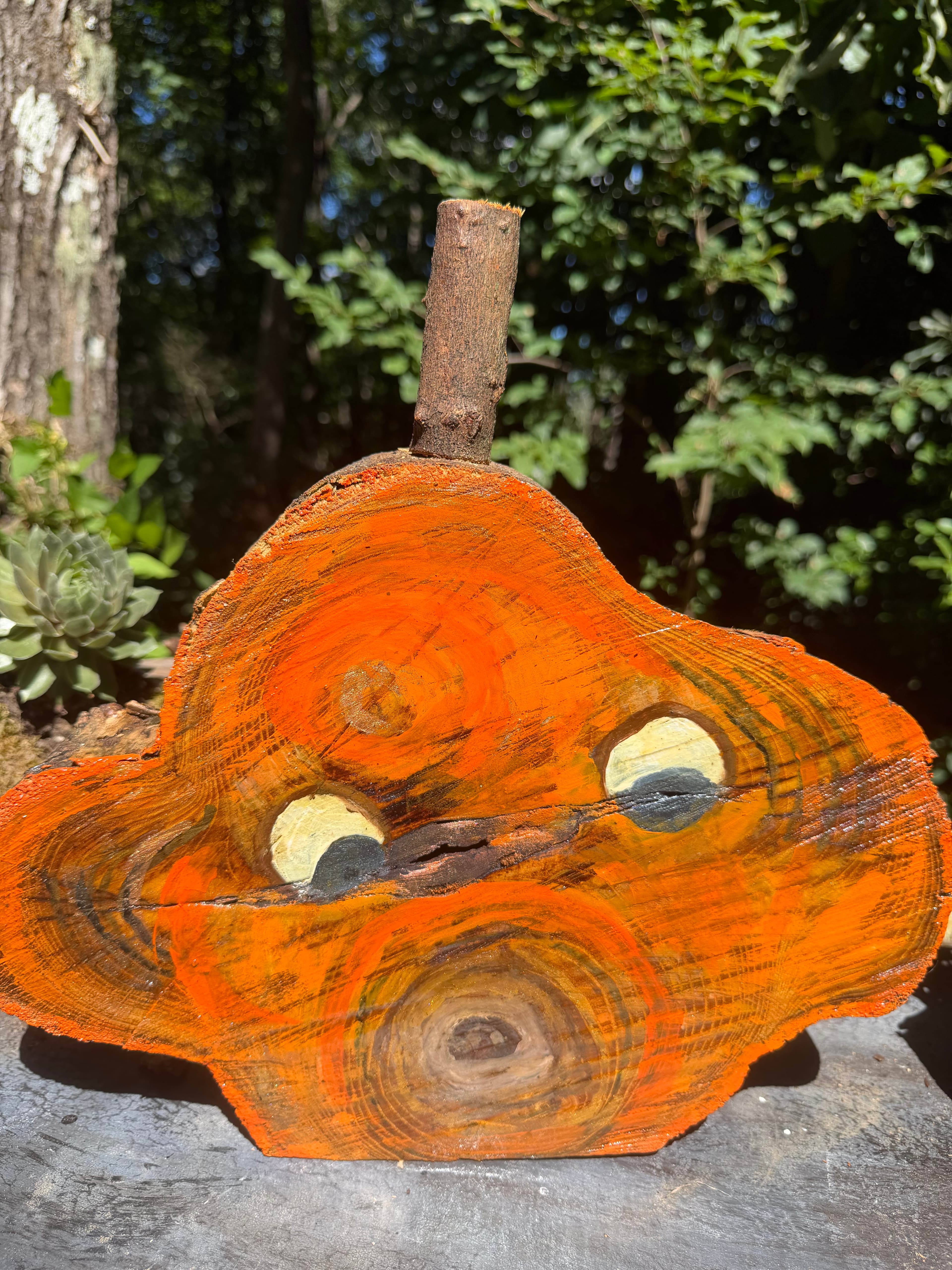 Rustic orange-painted wood slice pumpkin with a branch stem and painted eyes outdoors.